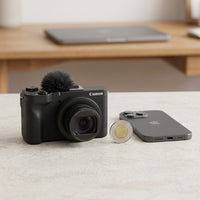 Black Canon V1 Vlogging compact camera with a furry microphone windscreen, shown on a stone surface beside a Canadian two-dollar coin and a smartphone for size comparison.