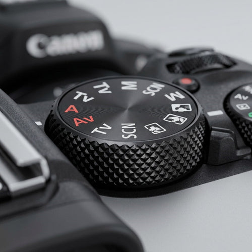 Close-up of the top mode dial on a black Canon EOS R50 camera, showing shooting-mode settings including M, Av, Tv, and SCN, with a textured knurled grip.