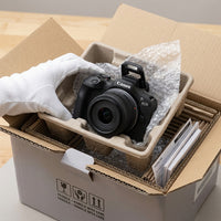 Close-up photo of the Canon EOS R100 camera and lens in protective cardboard inserts, being lifted from a clean shipping box.
