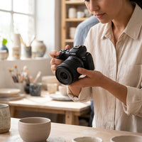 A woman holds a black Canon EOS R50 camera in a sunlit art studio, capturing photos of pottery.