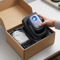 Hand lifting a Dangbei N2 Mini portable projector out of its foam-lined kraft retail box, with a white drawstring pouch and a coiled cable seated in the packaging beside it.