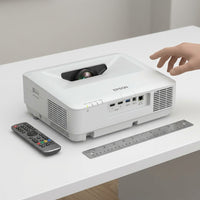 Epson BrightLink 770Fi projector shown to scale next to a standard AA-size remote control and a human hand on a white desk.