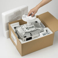 White Epson 3LCD projector packed in its box with a ceiling mount bracket attached, foam padding, a hardware bag of screws, and a manual; a hand reaches in to lift it out.