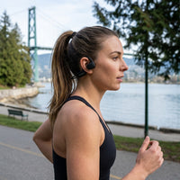 Woman in a black tank top running along a waterfront path, wearing black OPN Sound Aperto open-ear headphones, with a suspension bridge across the water behind her.