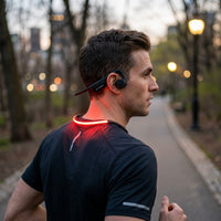 Man running on a tree-lined city path at dusk, wearing black OPN Sound Dashlyte+ open-ear headphones with a red illuminated LED safety collar.