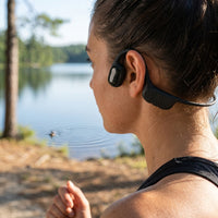 A sporty woman runs on a trail past a ripple on a lake, wearing black bone conduction headphones in natural light.