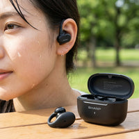 Woman in a green park wearing a black Sharp HP-TW45-BK open-ear true wireless earbud, with the second earbud and its open black charging case resting on a wooden table in front of her.
