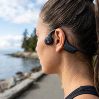 Runner wearing black Sharp HP-BC55 bone conduction headphones on a waterfront path, water droplets beaded on the ear hook