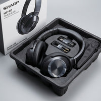 Black Sharp HP-60 over-ear wireless headphones seated in their moulded packaging tray, with a USB-C charging cable, a 3.5mm AUX cable, and a drawstring pouch, beside the retail box.