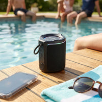 Black Sharp GX-BT190-BK portable Bluetooth speaker with water droplets on it, sitting on a wooden poolside deck beside a phone, sunglasses, and a striped towel, with people relaxing by the pool behind it.
