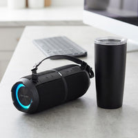 A photorealistic desk scene showing a SHARP GX-BT390 speaker beside a standard black travel tumbler for scale.