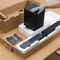 Sharp HT-SBW53121 soundbar and subwoofer unboxing: a gloved hand lifts the black subwoofer from its foam packaging, with the Dolby Atmos soundbar seated in the tray below and a remote and cables bagged alongside.