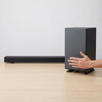 A Sharp HT-SBW53121 3.1.2 soundbar and wireless subwoofer are compared for size next to a human hand on a wooden desk.